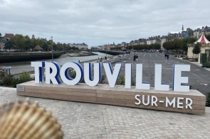 Duży biało-niebieski napis „TROUVILLE SUR-MER” stoi na drewnianym postumencie przy nadmorskim bulwarze. W tle widać rzekę wpływającą do morza, rząd kamienic i drzewa wzdłuż promenady. Kilka osób spaceruje po szerokiej ulicy, a niebo jest pochmurne.