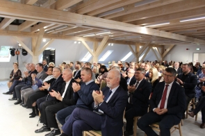 Duża grupa elegancko ubranych osób siedzi w jasnej sali z drewnianymi belkami na suficie i bije brawo. W pierwszym rzędzie znajdują się mężczyźni w garniturach oraz kobieta w czarnej sukni z białym koronkowym kołnierzem. Wszyscy patrzą w stronę sceny z uśmiechem, tworząc atmosferę uznania i uroczystości.