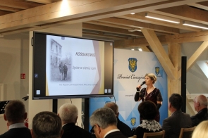 Kobieta w eleganckiej bordowej sukni z cekinami i długich czarnych rękawiczkach prowadzi prezentację, trzymając mikrofon. Na dużym ekranie za nią widnieje slajd z tytułem **„Kossakowie – życie w cieniu ruin”** oraz archiwalne zdjęcie. W tle widać logo **Powiat Cieszyński**, a przed sceną siedzi publiczność, skupiona na wystąpieniu. Całość odbywa się w nowoczesnym, jasnym wnętrzu z drewnianymi belkami i profesjonalnym sprzętem multimedialnym.