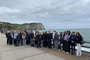 Grupa młodych osób stoi na nadmorskim deptaku z widokiem na morze i wysokie, białe klify z charakterystycznym łukiem skalnym w tle. Wiatr rozwiewa im włosy i ubrania, a niebo jest szare i pochmurne. Ludzie uśmiechają się i stoją blisko siebie, tworząc zwartą grupę.