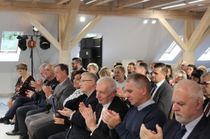 Grupa elegancko ubranych osób siedzi w jasnej sali z drewnianymi belkami na suficie i bije brawo. W pierwszym rzędzie widać mężczyzn w garniturach oraz kobiety w formalnych strojach. W tle znajdują się kolejne rzędy uczestników, którzy również uśmiechają się i klaszczą, tworząc atmosferę uroczystego wydarzenia.