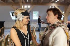 Dwie kobiety stoją naprzeciw siebie i rozmawiają w jasnym pomieszczeniu z drewnianymi belkami. Jedna z nich ma na sobie czarną sukienkę, długi naszyjnik z pereł i ozdobną opaskę z piórami na głowie, druga ubrana jest w jasny sweter i kolorową chustę w kwiaty. W tle widać rząd krzeseł oraz osobę niosącą tacę z poczęstunkiem.