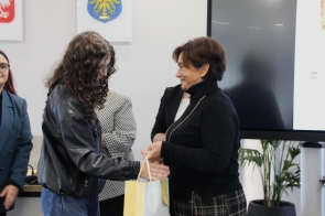 Janina Żagan ubrana na czarno, wręcza torbę z nagrodą laureatce I miejsca, ubranej w czarną kurtkę i niebieskie jeansy, włosy ma rozpuszczone kręcone.