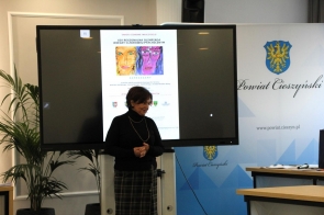 Janina Żagan Starosta Cieszyński ubrana w ciemny strój, stoi przed dużym ekranem w sali sesyjnej. Na ekranie widoczny jest slajd z kolorową grafiką i tekstem - plakat olimpiady. W tle znajdują się elementy dekoracyjne i fragment loga Powiatu na ścianie.