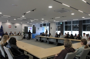 Uczniowie siedzą przy dużych stołach ustawionych w kształcie litery U w sali sesyjnej Starostwa Powiatowego w Cieszynie. Słuchają osoby stojącej przy ekranie z plakatem olimpiady. W tle widoczne są okna i elementy wyposażenia sali.