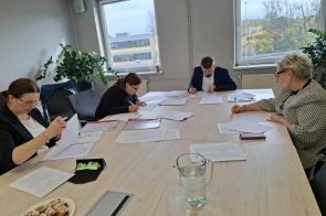 Jury  cztery osoby siedzące przy dużym stole w jasnym pomieszczeniu biurowym. Na stole znajdują się testy, długopisy, dzbanek z wodą. W tle są okna i rośliny.