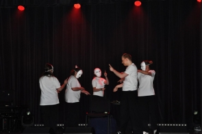 Grupa osób w białych koszulkach i białych maskach występuje na scenie oświetlonej czerwonym światłem, stojąc w półokręgu na tle ciemnej kurtyny.