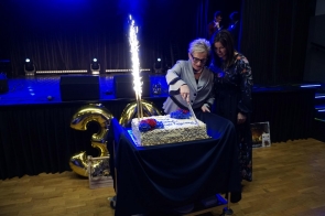 Kobieta w eleganckim, jasnoszarym kostiumie kroi duży tort ozdobiony czerwonymi i niebieskimi kwiatami oraz iskrzącymi fontannami; obok niej stoi druga kobieta w ciemnej sukni w kwiaty, a w tle sceny widać niebieskie światła i złote balony w kształcie liczby 30.