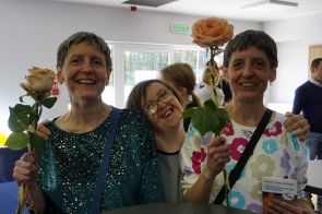 Trzy uśmiechnięte kobiety stoją blisko siebie w jasnym pomieszczeniu; dwie z nich trzymają w dłoniach pojedyncze róże, a środkowa obejmuje je ramionami.