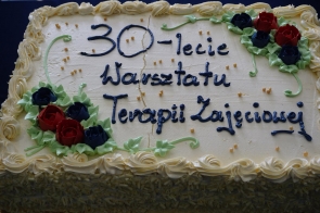 Prostokątny tort z jasnym kremem ozdobiony napisem „30-lecie Warsztatu Terapii Zajęciowej”, udekorowany kremowymi rozetkami na brzegach oraz kwiatami z czerwonego i granatowego lukru po bokach.