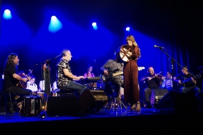 Na scenie oświetlonej niebieskim światłem występuje zespół muzyczny; kobieta w długiej bordowej sukni gra na bębnie obręczowym, obok niej muzycy grają na instrumentach takich jak gitara, skrzypce, keyboard i akordeon, a w tle siedzą uczestnicy warsztatu ubrani w białe koszule.