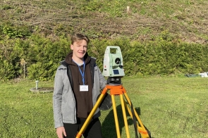 Młody mężczyzna stoi na zielonej łące obok sprzętu geodezyjnego ustawionego na statywie. Ma na sobie sportowe ubranie i identyfikator, a w tle widać porośnięte drzewami zbocze.