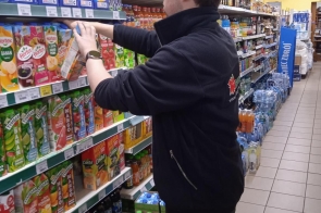 Młody mężczyzna w czarnej polarowej bluzie układa kartony soków na sklepowej półce. Znajduje się w alejce supermarketu, otoczonej napojami i wodami mineralnymi.