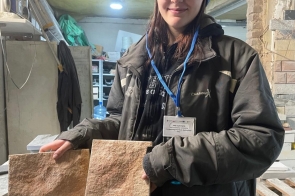 Młoda kobieta w roboczej kurtce, czapce i okularach ochronnych stoi w warsztacie, trzymając w dłoniach dwa prostokątne kamienne płytki. W tle widoczne są narzędzia, materiały budowlane i półki.