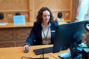 Młoda dziewczyna w eleganckim stroju biznesowym siedzi za recepcyjnym biurkiem hotelowym, pracując przy komputerze. Na biurku znajdują się monitor, tablet z logo hotelu Aries oraz terminal płatniczy.