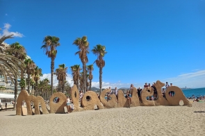 Grupa osób pozuje przy charakterystycznym napisie z piasku „Malagueta” na słynnej plaży w Maladze. W tle widać palmy, nadmorskie budynki i błękitne niebo, a po prawej stronie rozciąga się morze.