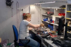 Młody mężczyzna siedzi przy biurku w warsztacie elektronicznym, skupiony na naprawie lub montażu urządzenia. Na stole leżą narzędzia, przewody i części elektroniczne, a obok stoi monitor komputerowy.