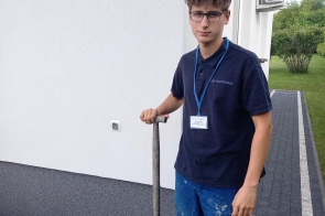 Młody mężczyzna w roboczym ubraniu stoi przed budynkiem, trzymając łopatę. Ma na sobie niebieskie spodnie z plamami farby i granatową koszulkę z identyfikatorem. Tło z zadbanym trawnikiem i białą elewacją.