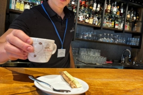 Młody mężczyzna w czarnej koszulce z identyfikatorem stoi za ladą w eleganckim barze. Z uśmiechem podaje filiżankę kawy, a na blacie leży talerzyk z łyżeczką i saszetką cukru. W tle widać półki z butelkami alkoholi i szkłem.