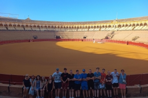 Grupa osób pozuje na tle dużego, okrągłego stadionu z piaskową areną i kamiennymi trybunami. Uczestnicy stoją w rzędzie przy barierce, uśmiechając się do zdjęcia w słoneczny dzień.
