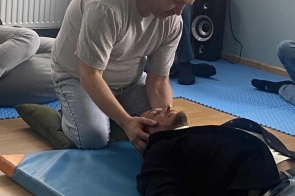 Mężczyzna w szarej koszulce klęczy na niebieskiej macie i delikatnie podtrzymuje głowę drugiego mężczyzny, który leży na plecach z rękami ułożonymi wzdłuż ciała. W tle widać kilku uczestników siedzących na matach, a za nimi kaloryfer i głośnik stojący przy ścianie.