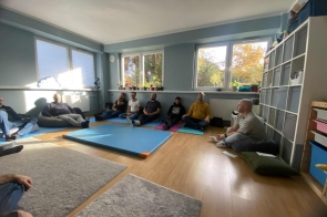 W niebieskiej sali grupa mężczyzn siedzi w kręgu na matach i poduszkach. Przed nimi leży niebieska mata, a przez duże okna wpada światło słoneczne, oświetlając pomieszczenie. Na parapetach stoją doniczki z roślinami, a po prawej stronie znajduje się regał z pudełkami i materiałami.