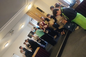 Grupa młodych osób odbiera dyplomy i upominki podczas uroczystości konkursowej. Uczestnicy stoją w dwóch rzędach, rozmawiają i przeglądają otrzymane nagrody, a w tle widoczne są banery organizatorów i sztandar szkoły.