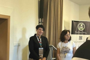 Dwie kobiety stoją obok siebie w pomieszczeniu, trzymając czerwone torby upominkowe. Jedna ma na sobie czarny żakiet i wzorzystą spódnicę, druga białą koszulkę z nadrukiem. W tle widać zasłonę, mównicę i baner z logo „Café.