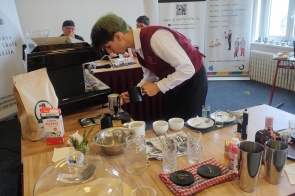 Młody chłopak w bordowym gorsecie i białej koszuli przygotowuje napój przy ekspresie do kawy, pochylony nad dzbankiem z mlekiem. Na stole przed nim stoją filiżanki, szklanki, shakery i różne składniki, a w tle widać jury obserwujące jego pracę.