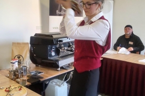 Młoda dziewczyna w bordowym gorsecie i białej koszuli energicznie potrząsa shakerem barmańskim, przygotowując napój. Ma związanego w kok włosy i nosi okulary, stoi przy stole z ekspresem do kawy, a w tle siedzi mężczyzna obserwujący jej pracę.