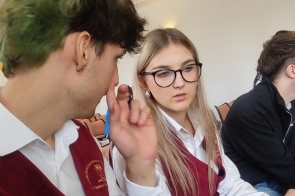 Dwie osoby siedzą obok siebie w sali, rozmawiając w trakcie wydarzenia. Oboje mają bordowe kamizelki z emblematem szkoły i białe koszule, a w tle widać innych uczestników oraz rząd krzeseł.