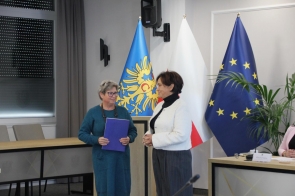 Po prawej stronie Janina Żagan Starosta Cieszyński, ubrana w białą marynarkę i ciemne spodnie, podczas wręczania zaświadczenia Marii Greń stojącej po lewej stronie w zielonej sukience. W tle widoczne flagi.