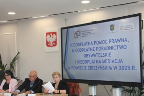 Trzy osoby siedzą przy stole prezydialnym w sali sesyjnej, przed dużym ekranem, na którym widnieje tytuł prezentacji dotyczącej nieodpłatnej pomocy prawnej, poradnictwa obywatelskiego i mediacji w powiecie cieszyńskim w 2025 roku. W tle widoczne są flagi. Otoczenie jest jasne i nowoczesne.