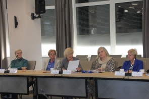 Pięć kobiet siedzi przy stole sesyjnym w sali obrad. Wszystkie są elegancko ubrane, przed nimi leżą dokumenty i mikrofony. Jedna z kobiet, siedząca pośrodku, czyta z kartki. W tle widać duże okna z zasłoniętymi roletami i herbem na szybach.