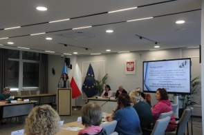 Kobieta stojąca przy mównicy prowadzi prezentację w nowoczesnej sali sesyjnej. Na ekranie obok wyświetlony jest slajd z informacjami dotyczącymi nieodpłatnej pomocy prawnej. Uczestnicy siedzą przy stołach.