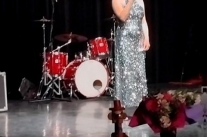 Elegancko ubrana młoda kobieta w błyszczącej, srebrnej sukni wykonuje utwór na scenie, a w tle widoczny jest czerwony zestaw perkusyjny oraz fragment baneru wydarzenia.