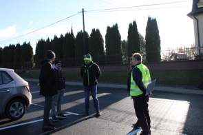 Na środku jezdni stoi grupa czterech osób rozmawiających ze sobą, po prawej jeden z nich ma na sobie jaskrawą kamizelkę odblaskową i trzyma dokumenty, a po lewej obok zaparkowany jest samochód. W tle widać rząd wysokich tuj rosnących za ogrodzeniem.