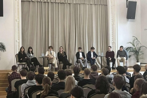 W jasnej sali ze sceną ustawiony jest rząd krzeseł, na których siedzi osiem osób. Przed sceną zgromadzona jest duża grupa młodych ludzi siedzących w kilku rzędach. W tle widać zasłony, a po bokach sceny stoją doniczkowe rośliny oraz stoliki z butelkami wody.