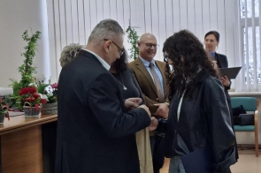 Na zdjęciu widać moment wręczania nagrody podczas uroczystości. Mężczyzna w garniturze przekazuje upominek młodej osobie o kręconych włosach, trzymającej teczkę. W tle stoją kolejne osoby obserwujące scenę, a obok widoczne są dekoracje kwiatowe ustawione na stole.