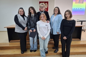 Na zdjęciu stoi sześć osób ustawionych w dwóch rzędach na tle sali konferencyjnej. W centralnej części zdjęcia stoi Beata Macura, Członkini Zarządu Powiatu Cieszyńskiego. W tle widać godło Polski oraz kolorowy slajd z prezentacji. Osoby pozują do wspólnej fotografii, uśmiechając się do obiektywu.