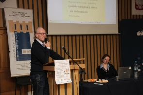 Mężczyzna stoi przy mównicy i mówi do mikrofonu, zwrócony w stronę publiczności. Obok niego znajduje się baner, a za plecami wyświetlona jest prezentacja. Po prawej stronie przy stole siedzi kobieta z laptopem, uśmiechając się lekko.