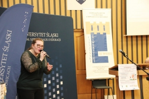 Młoda kobieta w okularach śpiewa do mikrofonu, gestykulując ręką. Stoi w jasnym, drewnianym pomieszczeniu obok dużych banerów i mównicy z mikrofonem.