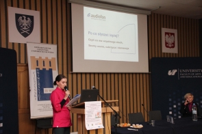 Kobieta w różowej, eleganckiej koszuli stoi przy mównicy, czytając z kartki i mówiąc do mikrofonu. Przed nią znajduje się laptop, a za nią wyświetlony jest slajd prezentacji dotyczący słuchu. W tle widać banery oraz drugą osobę siedzącą przy stole.