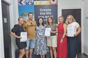 Sześć osób stoi razem w jasnym korytarzu na tle kolorowego plakatu. Trzy osoby trzymają certyfikaty. Wszyscy uśmiechają się i pozują do zdjęcia.