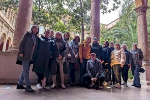 Grupa kilkunastu osób stoi razem w zadaszonym miejscu między kolumnami, a za nimi widać drzewa i rośliny. Wszyscy mają na sobie kurtki i pozują.