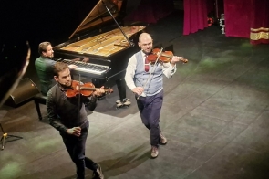Trzech muzyków występuje na scenie: dwóch gra na skrzypcach na przednim planie, a za nimi pianista siedzi przy dużym, otwartym fortepianie. Skrzypkowie poruszają się w rytmie utworu, a światła sceniczne delikatnie oświetlają ich sylwetki.