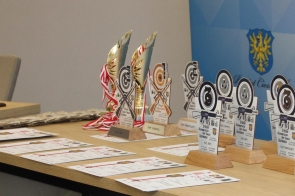 Stół wypełniony pucharami, statuetkami w kształcie tarcz strzeleckich i rozłożonymi dyplomami, w tle stoi ścianka z geometrycznym wzorem i herbem.