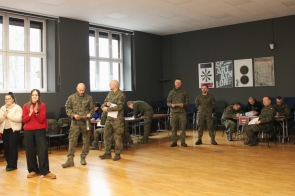 W jasnej sali z dużymi oknami stoi i siedzi grupa osób w wojskowych mundurach. Kilku żołnierzy trzyma dokumenty, inni zapisują się przy stołach ustawionych z tyłu sali. Po lewej stronie stoją dwie kobiety, w tym jedna w czerwonym swetrze, klaszcząca w kierunku grupy. Na ścianie wiszą plakaty graficzne, a podłoga jest drewniana.