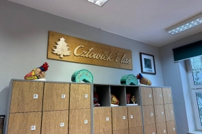 Drewniana tablica z napisem „Człowiek i las” oraz symbolem choinki wisi nad rzędem szafek. Na szafkach stoją kolorowe modele anatomiczne serc oraz kilka innych edukacyjnych figurek. Po prawej stronie znajduje się okno oraz obraz w ramie.