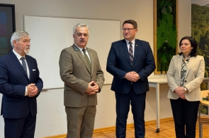 Cztery osoby stoją obok siebie w jasnym pomieszczeniu, ustawione w lekkim półokręgu. Dwaj mężczyźni po lewej mają na sobie eleganckie garnitury, a trzeci — drugi od lewej — nosi mundur w odcieniach zieleni. Po prawej stronie stoi kobieta w jasnej marynarce i wzorzystej koszuli. W tle widać białą tablicę oraz dekorację z roślin w ramie.